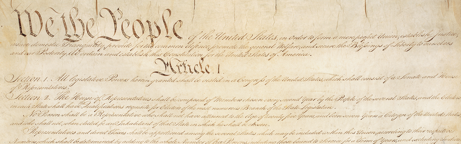 A cropped photograph of the United States Constitution on parchment paper and written in elaborate penmanship cursive writing.